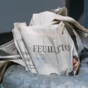 Eine gedruckte Zeitung steckt im Mülleimer einer Haltestelle. Aufgeschlagen ist das Feuilleton.