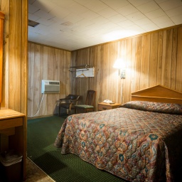 Ein Zimmer in einem günstigen Motel. Mit falscher Holzvertäfelung und grünem Teppich unter dem geblümten Bett.