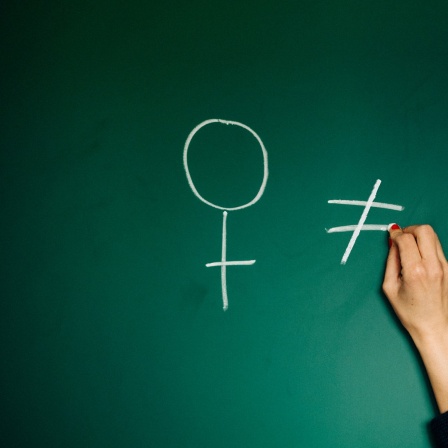 Eine Hand zeichnet die Gendersymbole an eine Tafel, in der Mitte ist ein "Ungleich"-Zeichen