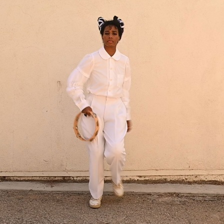 Santigold: "Shake" - die Sängerin, ganz in weiß gekleidet mit einem Tambourin in der Hand vor einer Wand stehend.