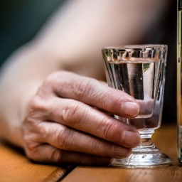Die Hand eines alten Mannes hält ein Schnapsglas. Der Arm an der Hand ist nackt. Neben dem Glas steht eine volle Glasflasche.