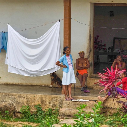 Vier kubanische Frauen halten sich vor einem Wohnhaus auf. Drei Frauen sitzen, eine steht. Ein Bettlaken hängt auf einer Wäscheleine. Im Hintergrund sitzt ein Hund. 