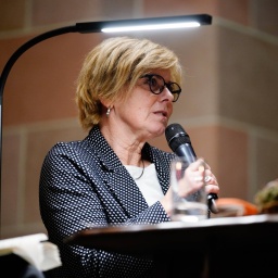 Verena Welschof sitzt bei einer Lesung an einem Schreibtisch. Sie trägt kurzes mittelblondes Haar und einen schwarzen Blazer mit kleinen weißen Pünktchen.