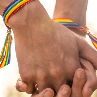 Zwei Hände greifen ineinander, an den Handgelenken sind Freundschaftsbänder mit den Regenbogenfarben festgebunden. (Symbolbild LGBTQ)