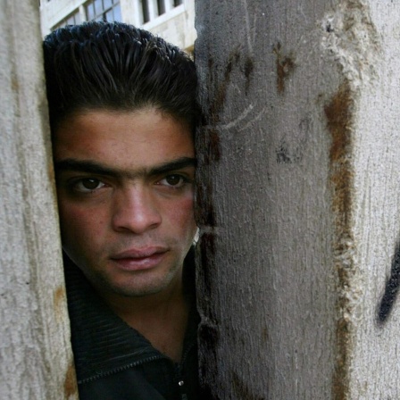 Ein Palästinenser, der auf der Westjordanlandseite des "Trennzauns" in Abu Dis steht, schaut im November 2003 durch eine Lücke, um zu sehen, ob israelische Polizisten in der Nähe auf der Ost-Jerusalem-Seite der Mauer sind. Auf der Mauer steht &#034;Apartheid&#034;.