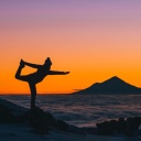 Eine junge Frau macht in Neuseeland Yogaübungen bei Sonnenuntergang über den Wolken, im Hintergrund ist der Mount Taranaki zu sehen.
      