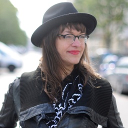 Die Comiczeichnerin und Illustratorin Ulli Lust mit Brille und Hut und schwarz gekleidet steht auf einer Straße, neben ihr parkende Autos.