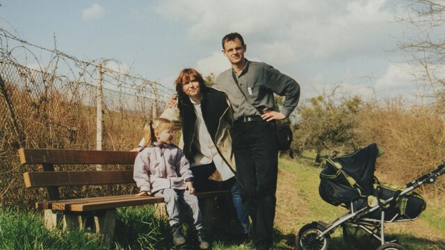 Szene aus Kurzzeitschwester: Auf einer Bank in der Natur aufgenommenes Familienfoto der Eltern des Autoren mit der kleinen Adoptivtochter.