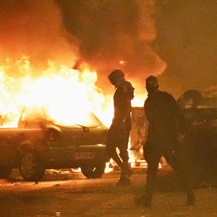 Demonstranten stehen in Paris auf einer Straße im Dunkeln, im Hintergrund sind brennende Fahrzeuge zu erkennen.