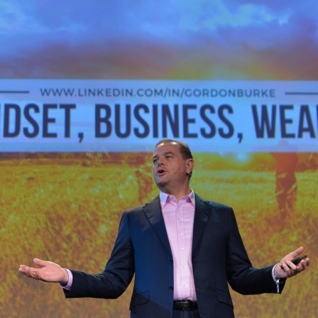 Ein Mann im Anzug steht mit ausgebreiteten Armen auf einer Bühne. Hinter ihm wird das Bild einer sonnenbestrahlten Wiese projiziert, darauf die Worte: "Mindset, Business, Wealth".
