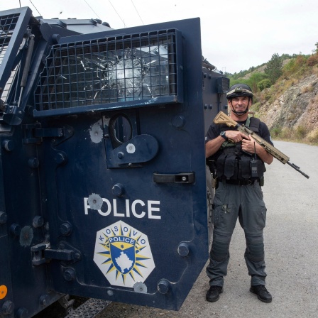 Ein Polizist steht im Kosovo neben einem Panzerwagen, er hält ein Sturmgewehr schräg vor der Brust und grinst