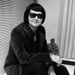 Der amerikanische Sänger Roy Orbison sitzt vor einer Jalousie und schaut durch seine dunkle Sonnenbrille.
