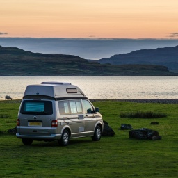 Im Abendlicht steht ein Campervan auf einer grünen Wiese zwischen Schafen direkt an einem idyllischen Platz am Meer. Grossbritannien.