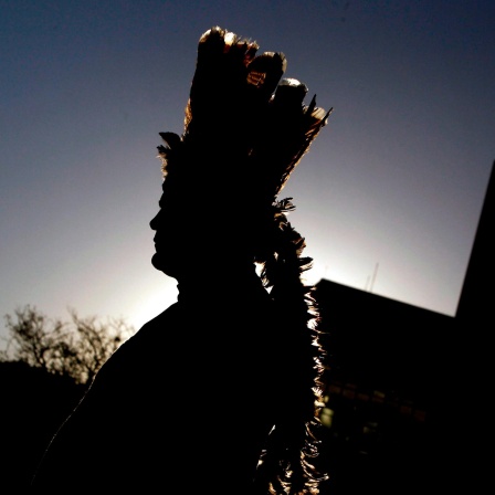 Die Silhouette eines Indigenen mit einem Federkopfschmuck hebt sich vor einem dämmerigen Himmel ab.