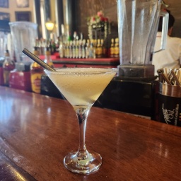 Ein Cocktailglas steht auf einer Bar, es enthält einen hellgelben Drink und einen Trinkhalm aus Metall.