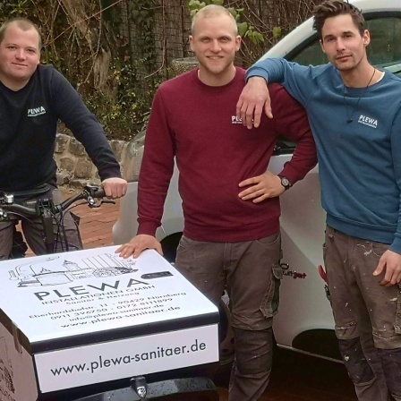3 junge Männer. Einer links auf einem Lastenfahrrad, 2 stehen rechts davon an ein Auto gelehnt.