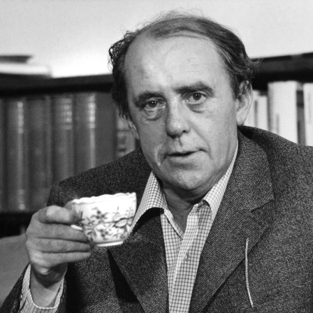 Schwarzweißaufnahme von Heinrich Böll, der mit Kaffeetasse und Zigarette in seiner Wohnung an einem Kaffeetisch sitzt.