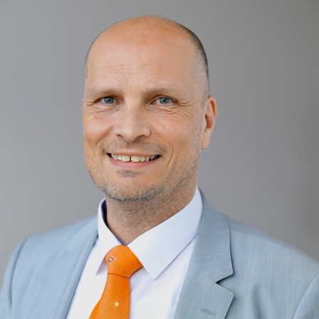 Ein Porträt von  Prof. Dominik Enste in Anzug und Krawatte vor grauem Hintergrund.