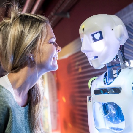 Eine Frau steht lächelnd vor einem Roboter, der Herzen in den Augen hat (Bild: picture alliance / Westend61)