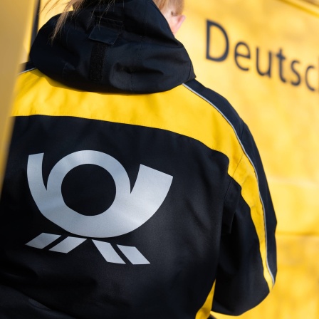 Eine Postbotin trägt vor einem Zustellfahrzeug eine Jacke mit dem Logo der Deutschen Post. R