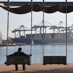 Zwei Personen sind als Silhouette zu sehen wie sie auf Bänken sitzend in Richtung eines Containerhafens mit einem großen Cargoschiff schauen.