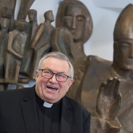 Kardinal Lehmann wird 80: Ein lachender Kardinal am Bildrand, im Hintergrund christliche Bronzestatuen.