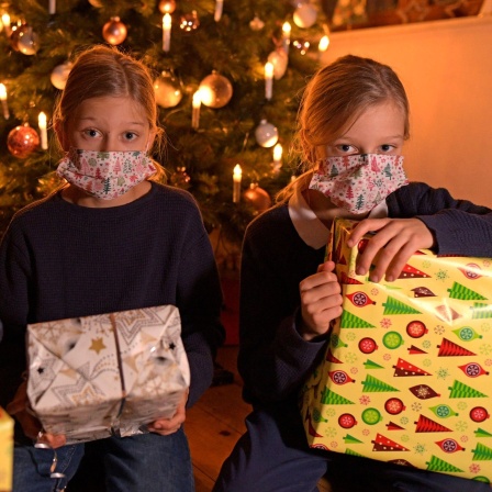 Drei Kinder mit Masken und Geschenken vor dem Weihnachtsbaum