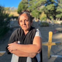 Eine Frau steht mit verschränkten Armen auf einem Friedhof, im Hintergrund sind Kreuze zu sehen