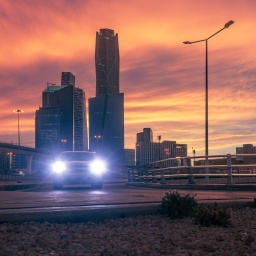 Skyline des Köng Abdullah Finanzdistrikt (KAFD) in Riyadh vor orangenem Himmel. Die Scheinwerfer eines Fahrzeugs leuchten in Richtung der Kamera.