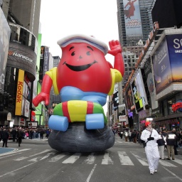 Das Bild zeigt ein riesiges aufgeblasenes rotes Maskottchen mit lachendem Gesicht, das winkt. Der große Ballon wird während einer Parade durch die Straßen von Manhattan gezogen. 