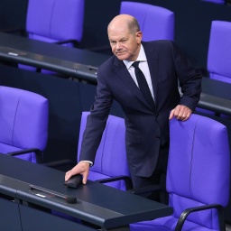 Bundeskanzler Olaf Scholz (SPD) hält sich an einem Stuhl auf der Regierungsbank fest und legt etwas ab.