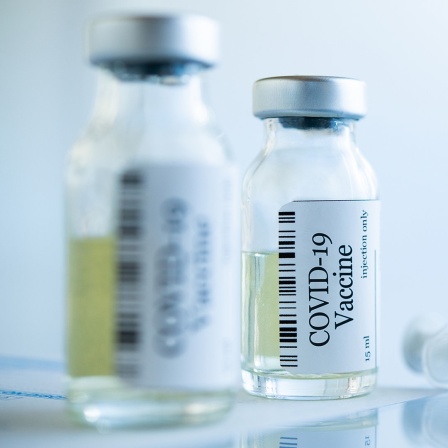 Zwei Fläschen mit Covid-19-Impfstoff und Spritzen liegen auf einem Glastisch. 