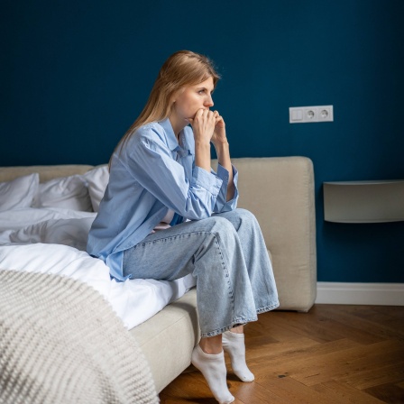 Eine besorgte Frau sitzt auf dem Bett, schaut aus dem Fenster und grübelt