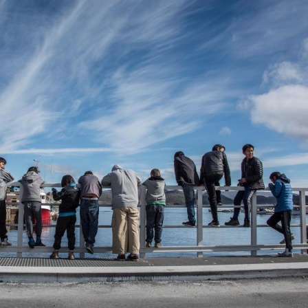 Eine Gruppe jugendlicher Jungs steht an einer hölzernen Uferbegrenzung. Manche sind daran gelehnt und schauen aufs Wasser, andere sitzen rittlings darauf.