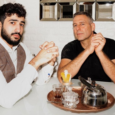 Mohamed Chahrour und Marcus Staiger, beide mit ineinander verschränkten Fingern, am Tisch sitzend.