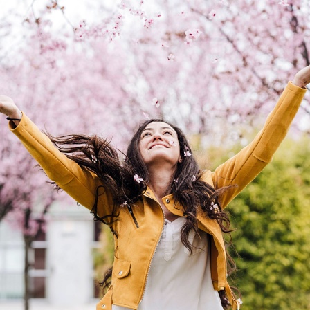 Junge Frau freut sich, mit einer leichten Übergangsjacke bekleidet, über die Kirschblüte im Frühling.