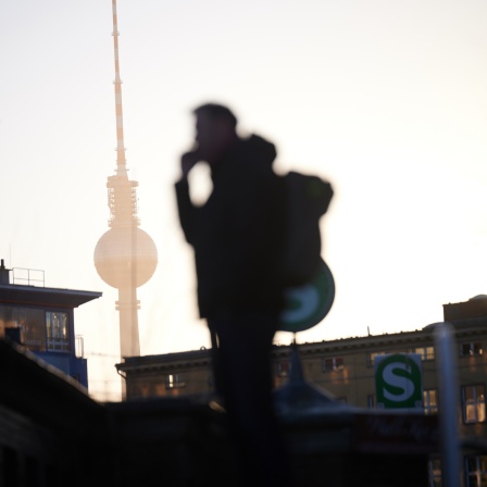 Die Sonne geht hier dem Fernsehturm auf, während ein Mann mit Rucksack am Nordbahnhof im Gegenlicht steht und telefoniert.