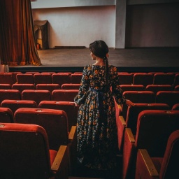Im Theatersaal: Eine elegante Frau im Kleid steht im Gang zwischen den leeren Zuschauersitzen und blickt nachdenklich gen Theaterbühne.