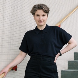 Annabelle von Reutern steht auf einer Treppe und hält sich entspannt mit einer Hand am Geländer fest – Sie hat kurzes, gräuliches, nach hinten gekämmtes Haar, braune Augen, helle Haut und trägt ein schwarzes Poloshirt.