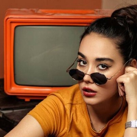 Frau mit heruntergeschobener Sonnenbrille auf der Nase sitzt vor einem orangenen Retro-Fernseher.