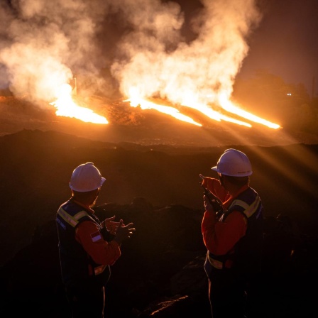 Nickelproduktion in Indonesien. Zwei Wachposten mit weißen Helmen und Feuerschutzwesten beobachten gleißendes Licht auf einer Mine. Das Licht ist in Orange getaucht.