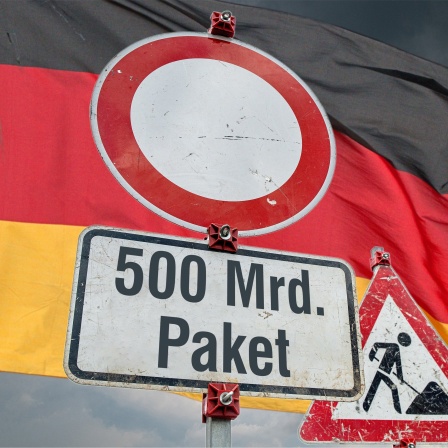  Schilder an einer imaginaeren Baustelle. Verkehrszeichen 250 (Verbot fuer Fahrzeuge aller Art) in runder Form, ein Zusatzschild mit der Aufschrift: "500 Mrd. Paket" an einem Schildermast und im Hintergrund das Schild Zeichen 123 Arbeitsstelle bzw. Baustelle vor einem grau bewoelkten Himmel mit aufziehendem Gewitter und einer, im Wind wehenden Nationaflagge der Bundesrepublik Deutschland. Foto mit Composing als Symbolbild fuer das, von Union (CDU / CSU), SPD und Gruenen beschlossene Sondervermoegen von 500 Milliarden Euro fuer zusaetzliche Investitionen in die Infrastruktur ueber 12 Jahre.