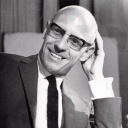 Schwarzweiße Porträtfotografie des Philosophen Michel Foucault: Er blickt in zugewandter Haltung in Richtung eines Gesprächspartners, lächelt dabei und fasst sich mit einer Hand an die Glatze.
