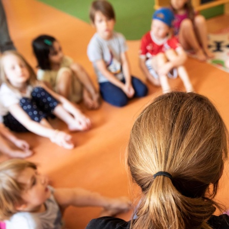 Eine Erzieherin sitzt mit Kindern im Kindergarten im Kreis auf dem Boden