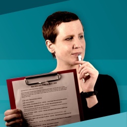 Eine junge Frau mit kurzen dunklen Haaren und schwarzem Shirt hält in ihrer rechten Hand ein Klemmbrett mit Zettel und in der linken einen Kugelschreiber, dessen oberes Ende ihre Unterlippe berührt.