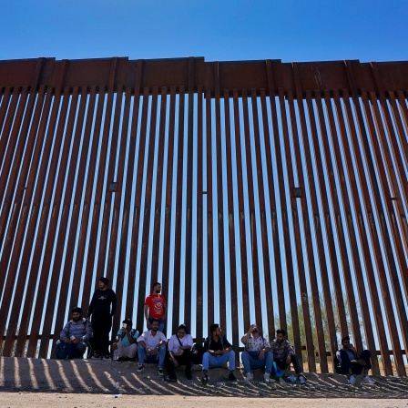 Das Beitragsbild des Dok5 "Auf Turnschuhen durch die Wüste" zeigt eine Gruppe Flüchtlinge im Schatten der amerikanischen Grenzmauer in der Region Tucson in 2023.