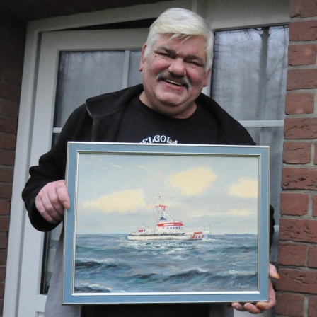 Ein Mann lächelt in die Kamera und hält ein Gemälde hoch, auf dem ein Seenotrettungsschiff zu sehen ist.