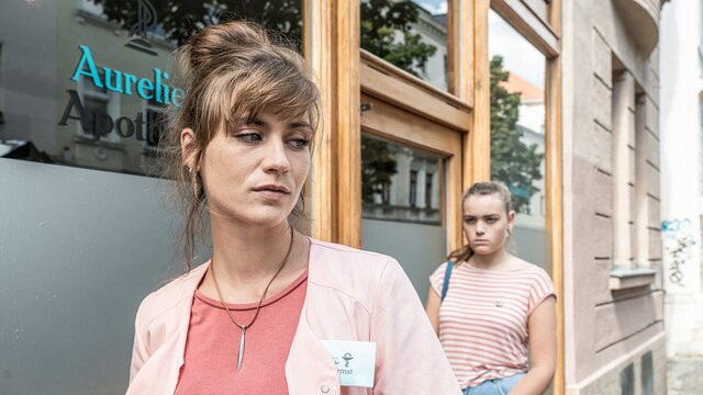 Sandra (Sophie Pfennigstorf) vor der Apotheke, in der sie arbeitet. Hinter ihr steht das Teenie-Mädchen Mia (Shenia Pitschmann).