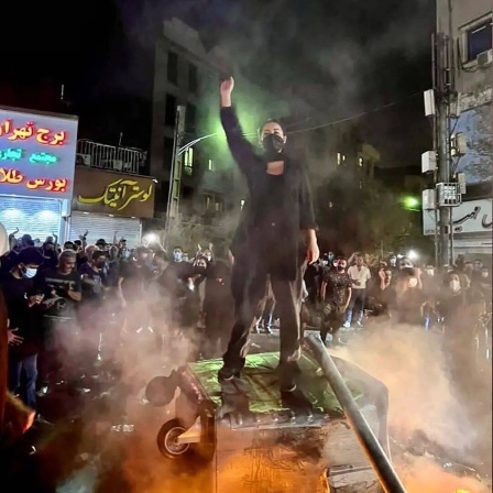 Eine Frau bei einer Demonstration im Iran, die auf einer großen Mülltonne steht und den Arm hebt