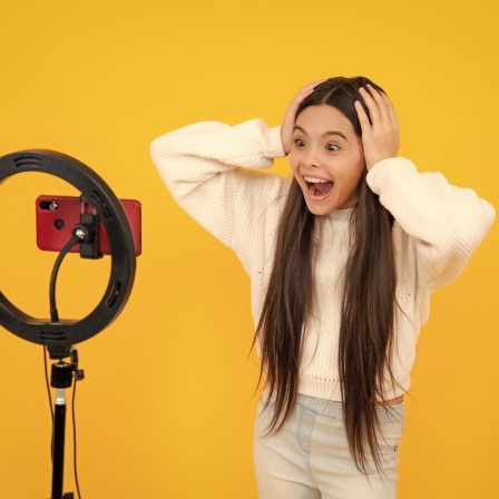 Eine junge Bloggerin vor dem Kameraphone mit weit aufgerissenen Augen vor gelbem Hintergrund.